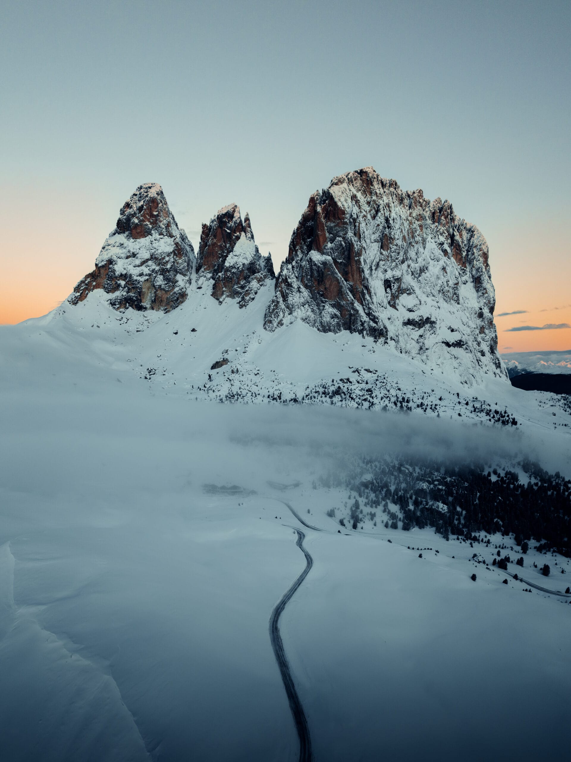 Snow-covered mountains rise above a misty landscape at dawn, with a winding road leading through the snow in the foreground—the perfect scene for an Outdoor Filmmaker capturing nature’s beauty beneath a subtle orange glow on the horizon.