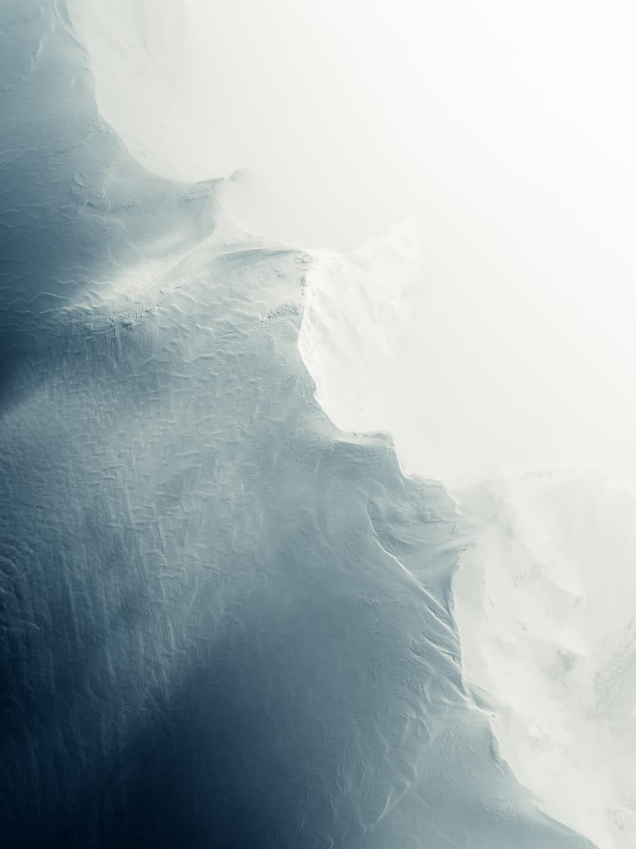 Aerial view of a snowy landscape with smooth, white snow merging into darker, textured terrain. Italian Content Creator Filippo Bellisola captures this cold and serene scene, showcasing gentle curves and subtle patterns in a soft gradient from light to dark.