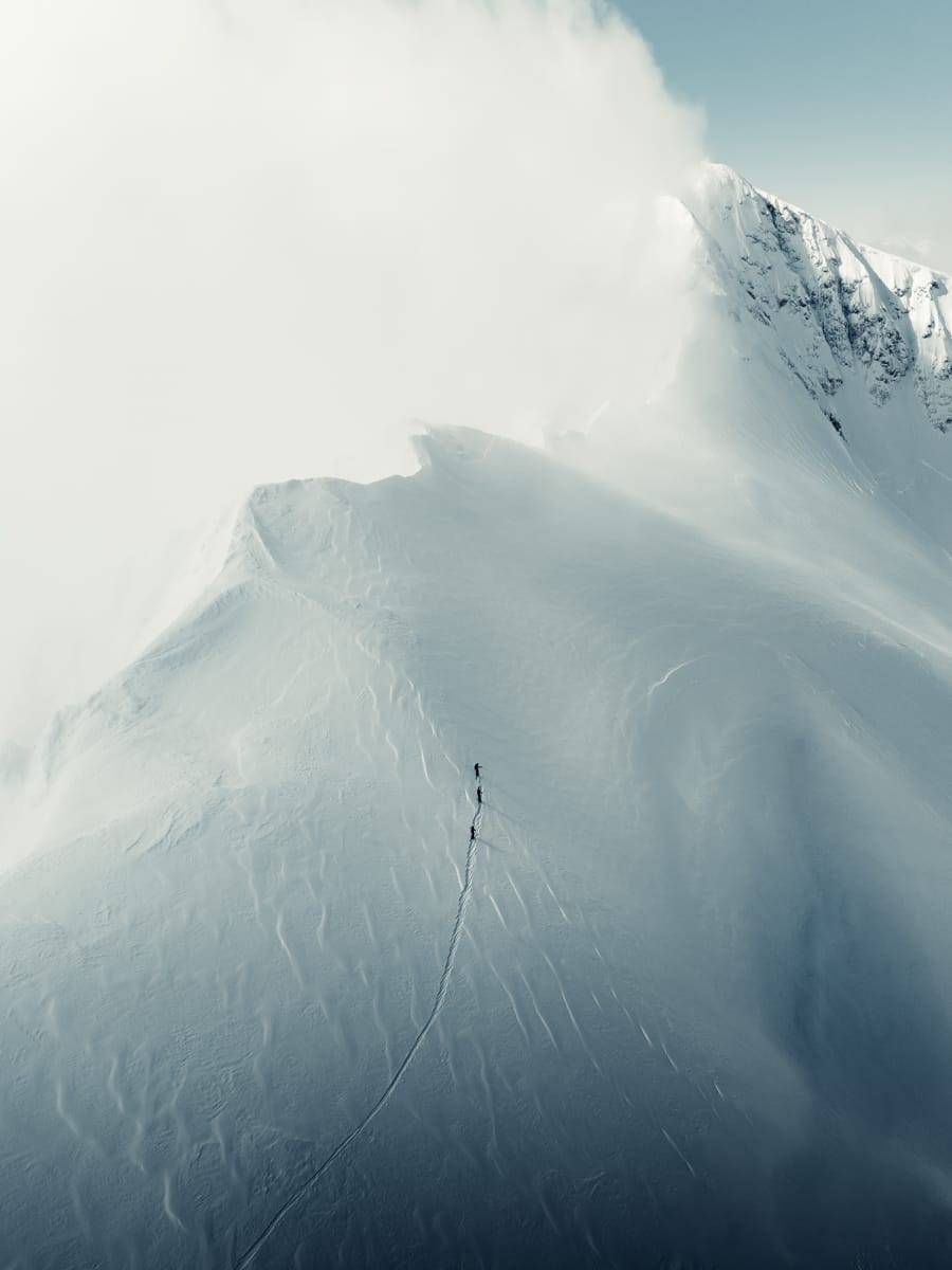 A lone skier ascends a snowy mountain ridge, leaving a single trail behind—an inspiring scene for any outdoor filmmaker, set against dramatic clouds and rugged peaks in the background.