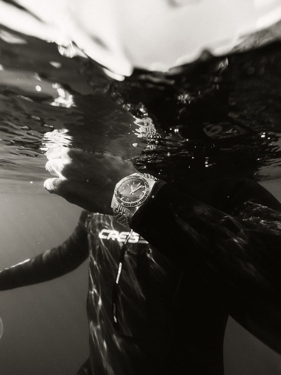 A person in a wetsuit, like Outdoor Content Creator Filippo Bellisola, is partially submerged underwater, their wristwatch glinting just below the surface as light reflects off both the watch and the rippling water.