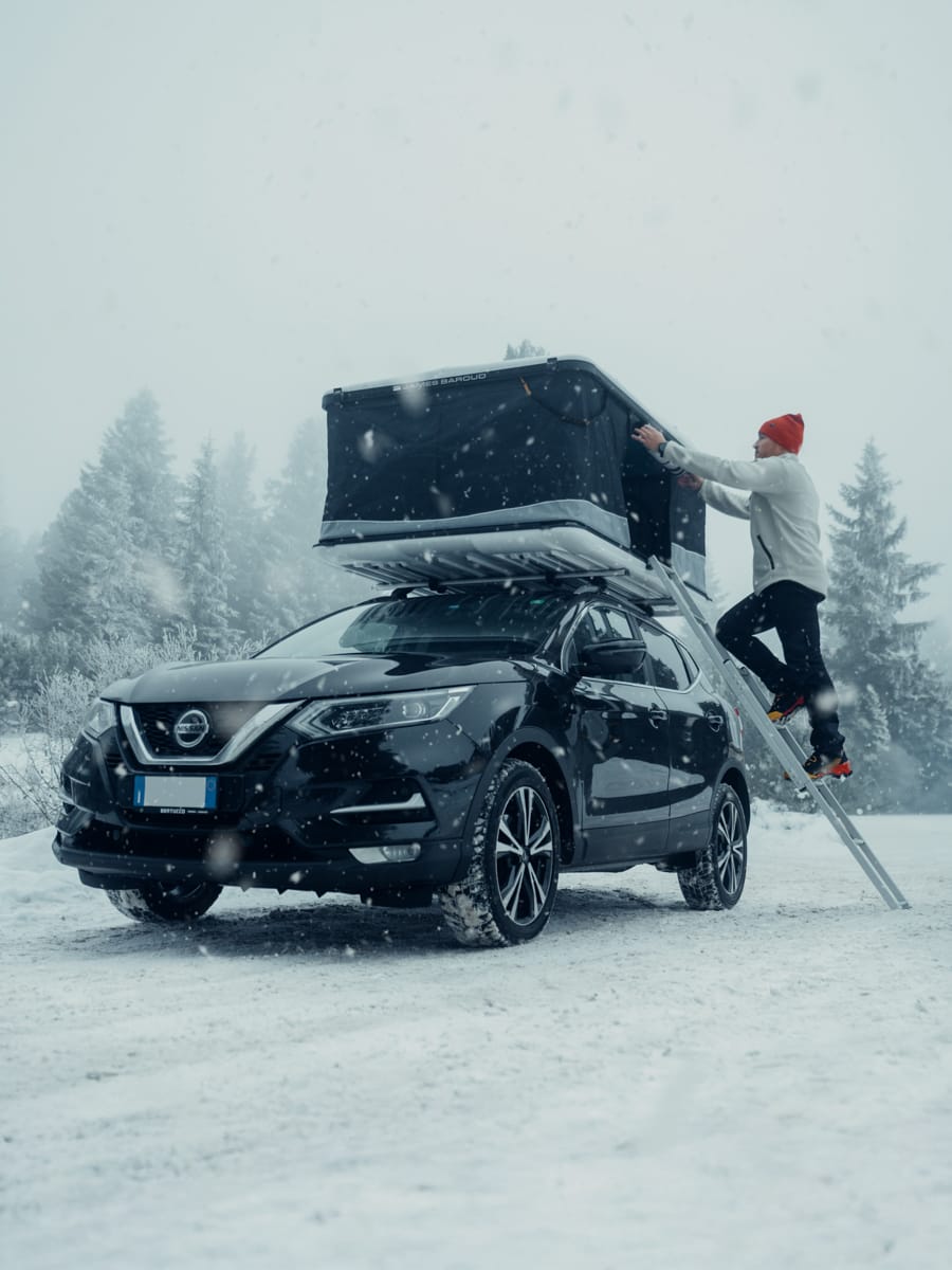 Outdoor Content Creator Filippo Bellisola climbs a ladder to a rooftop tent on a black SUV parked in a snowy, forested landscape. Snow is falling, capturing the cold and wintry adventure.