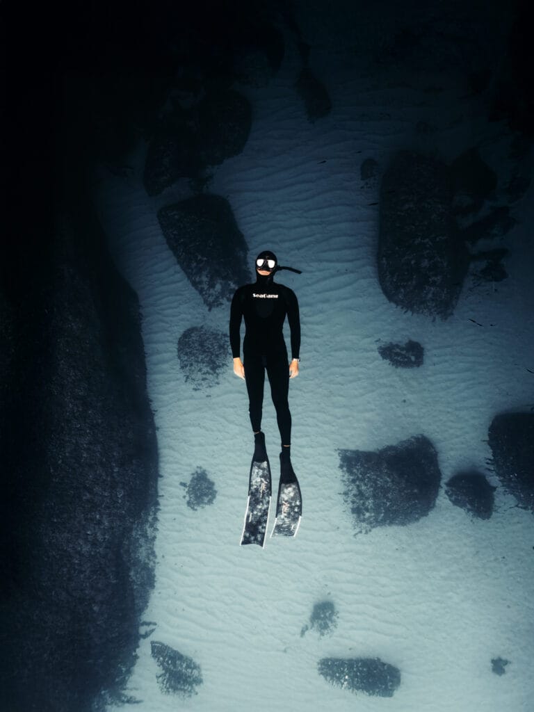 A diver in a black wetsuit, snorkel, and fins floats motionless underwater above a sandy, rocky seabed—a scene captured by Italian Content Creator and Outdoor Filmmaker Filippo Bellisola, surrounded by scattered rocks and dim natural light.