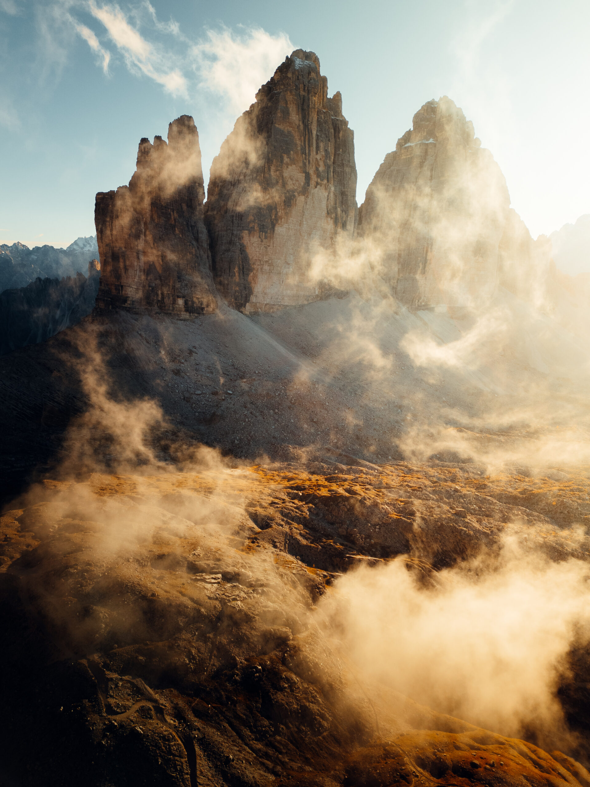 Three rugged mountain peaks rise dramatically above rocky terrain, with golden sunlight filtering through swirling mist and clouds—a striking, atmospheric landscape scene perfect for any outdoor content creator.