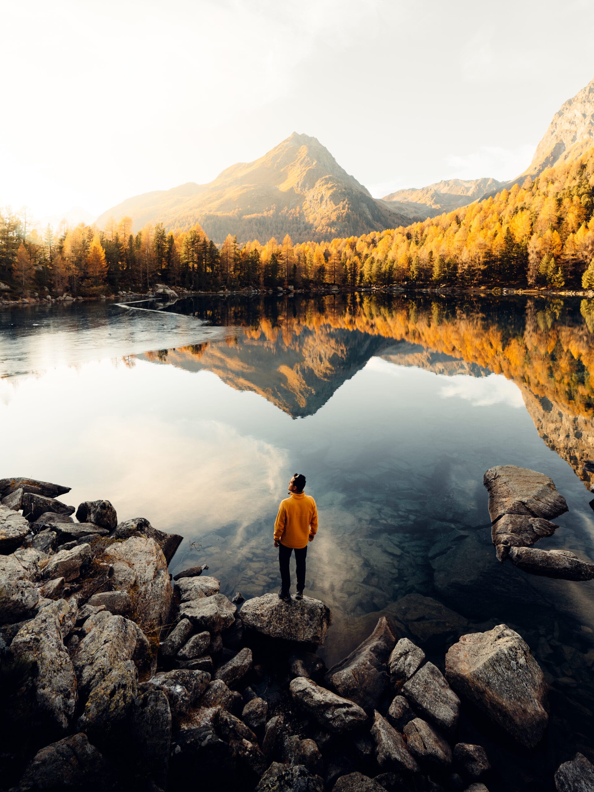 A person in a yellow jacket stands on a rock by a calm lake, surrounded by autumn trees. This serene moment, captured by Italian content creator Filippo Bellisola, reflects mountain peaks and trees under a bright, partly cloudy sky.