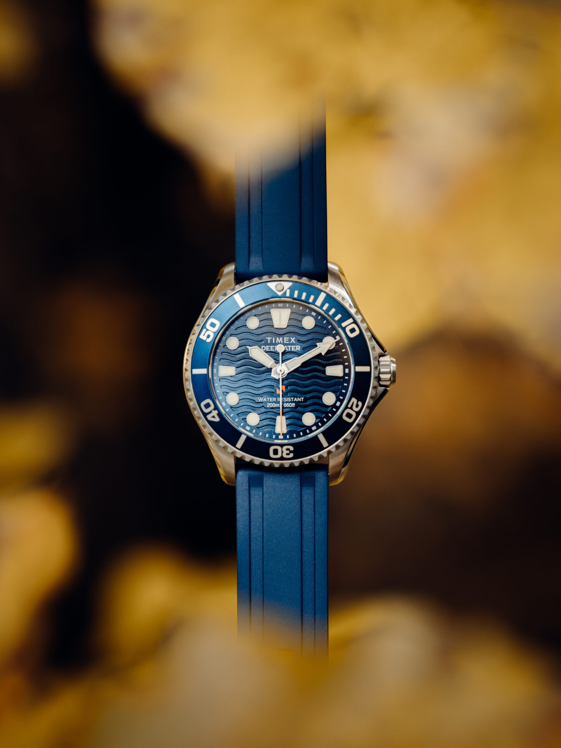A blue sports wristwatch with a wavy pattern on the dial and a blue strap, centred in the image—perfect for any Outdoor Content Creator—set against an out-of-focus, golden-brown background.