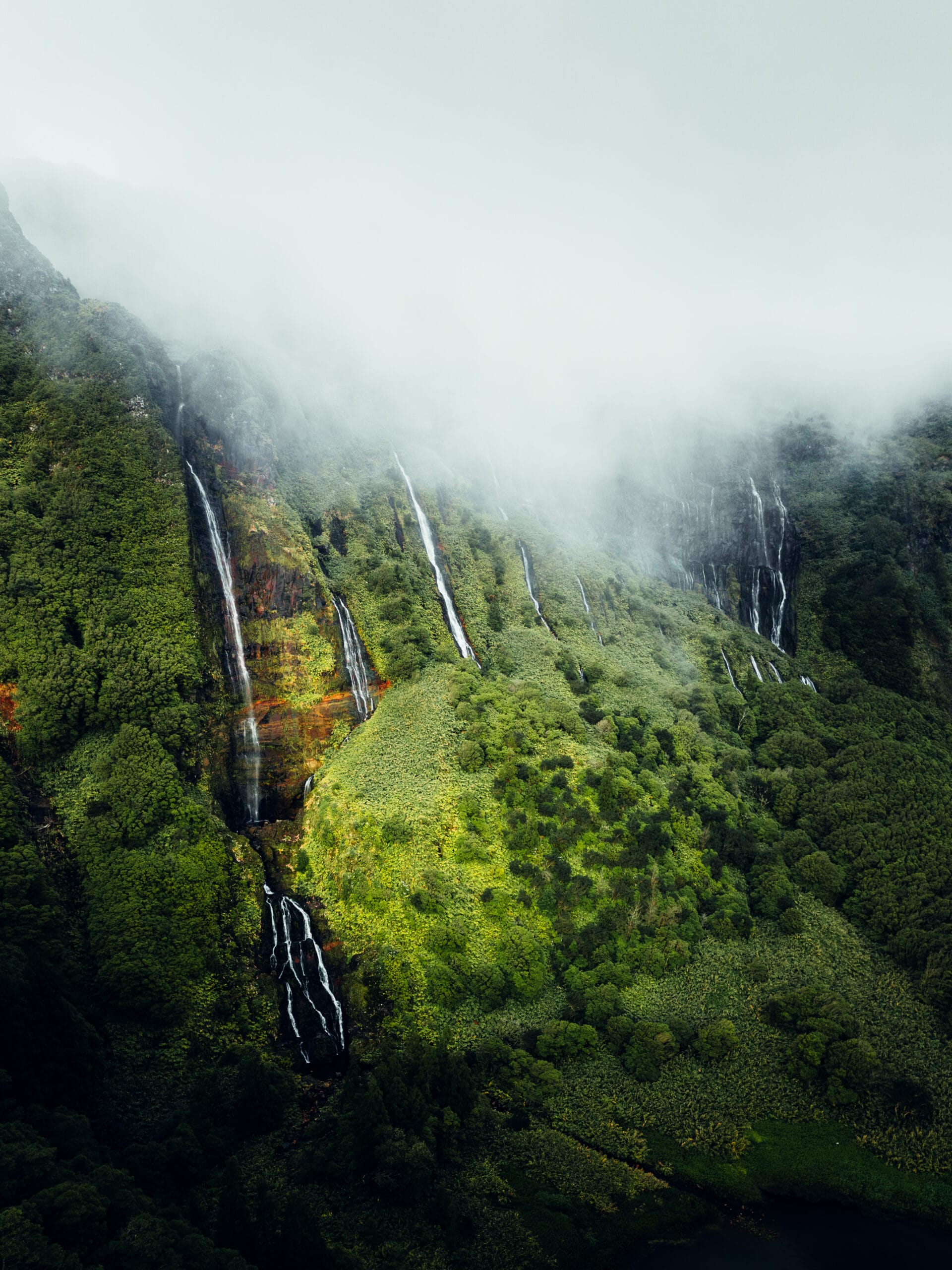 A lush green mountainside with several thin waterfalls cascading down, partially covered by mist and low-hanging clouds—an inspiring scene for any outdoor film-maker seeking breath-taking natural beauty.