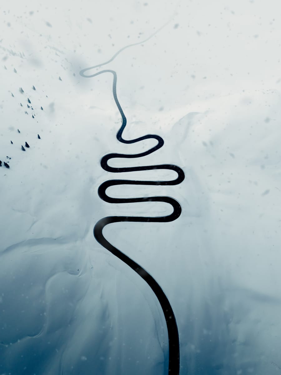 A winding, zigzag road cuts through a snowy, mountainous landscape, surrounded by white snow and scattered dark trees. This serene, minimalistic scene is perfect inspiration for any Outdoor Filmmaker seeking breathtaking aerial perspectives.