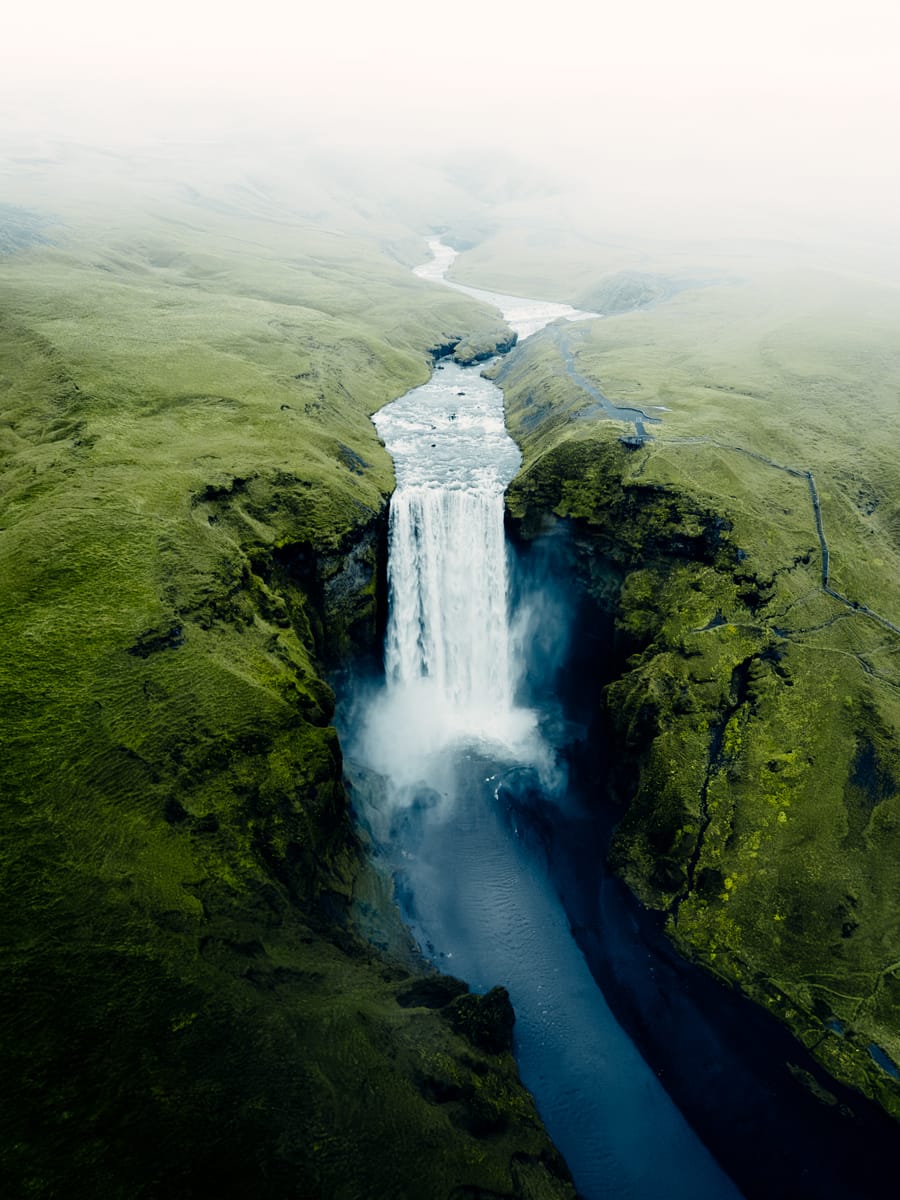 A tall waterfall cascades into a river, surrounded by lush green cliffs and mist—captured expertly by Outdoor Filmmaker Filippo Bellisola—with rolling hills fading into the distance under a pale sky.