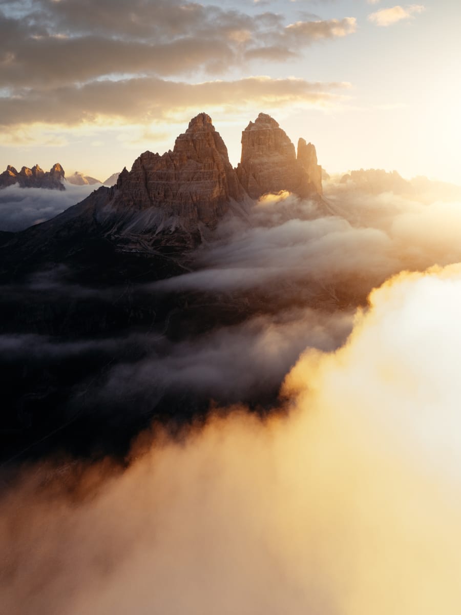 Sunrise over rugged mountain peaks, partially veiled by clouds and mist, with warm golden light illuminating the scene—an inspiring moment captured by Outdoor Filmmaker Filippo Bellisola.