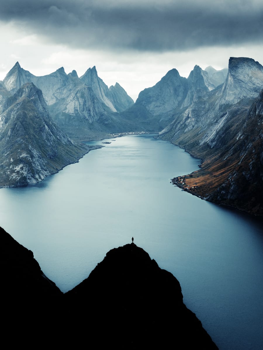 A person stands on a dark mountain peak overlooking a dramatic fjord surrounded by sharp, rugged mountains under a cloudy sky, capturing the moment like an outdoor film-maker inspired by Filippo Bellisola.