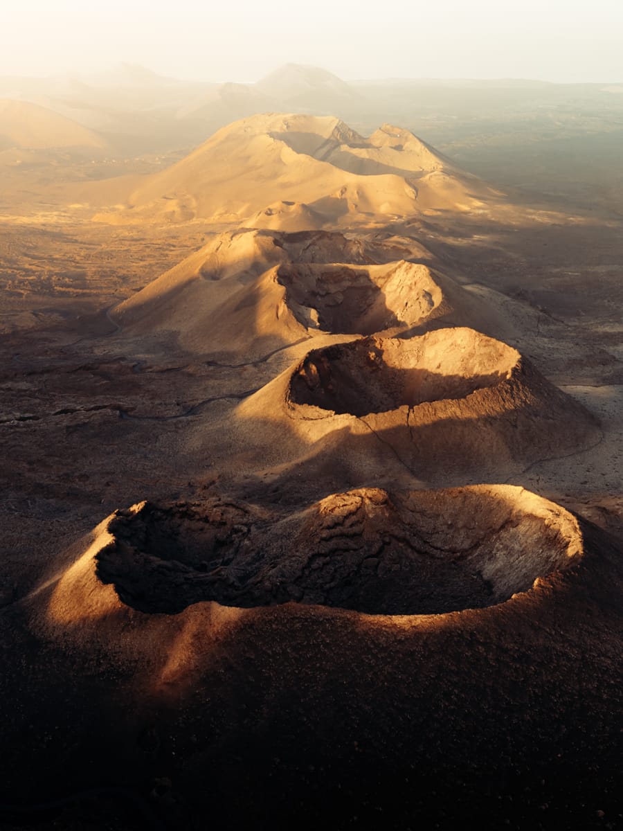 Aerial view of volcanic craters and rugged terrain, lit by golden sunlight, surrounded by arid landscape and hazy mountains—perfect inspiration for an outdoor film-maker capturing dramatic natural beauty.