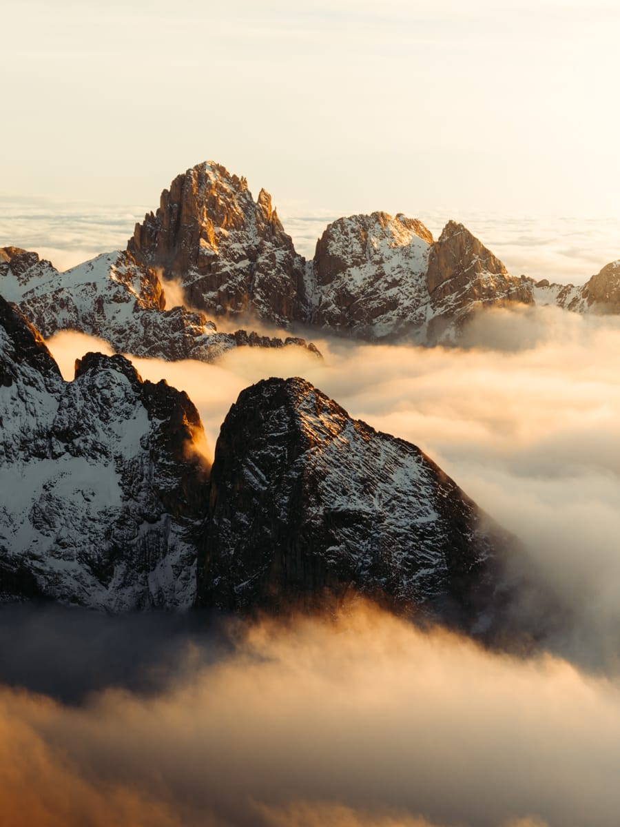 Snow-capped mountain peaks rise above a blanket of clouds at sunrise, with warm golden light illuminating the rocky summits and mist softly drifting through the valleys—a scene worthy of a Travel Filmmaker like Filippo Bellisola.