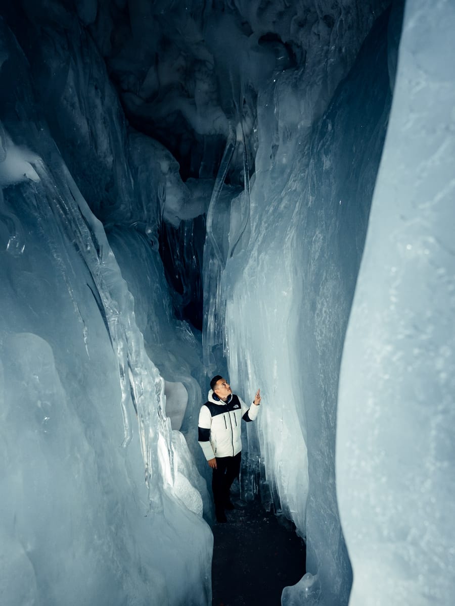 A travel filmmaker wearing a white and black jacket stands inside a narrow icy cave, touching the smooth, towering walls of blue-toned ice that surround them.