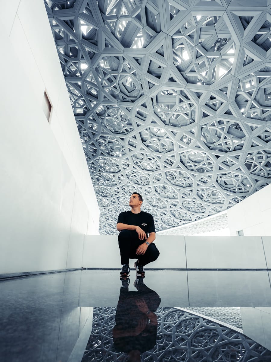 A person in black attire crouches on a shiny floor, gazing up at an intricate, geometric patterned ceiling reflected below—capturing the scene like an Italian Content Creator exploring striking modern architecture.