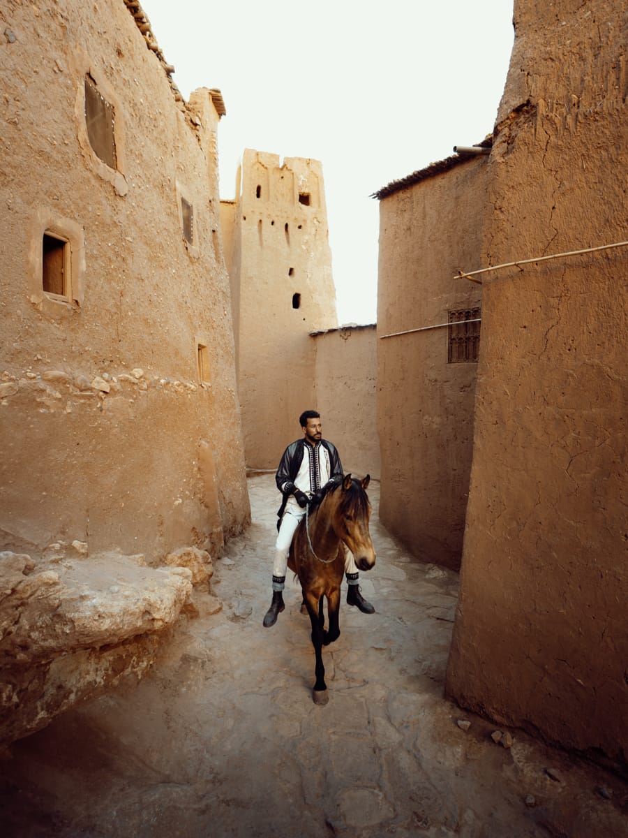 Travel Content Creator Filippo Bellisola, dressed in a white outfit and dark jacket, rides a brown horse through a narrow alleyway lined with tall, mud-brick buildings beneath a bright sky.