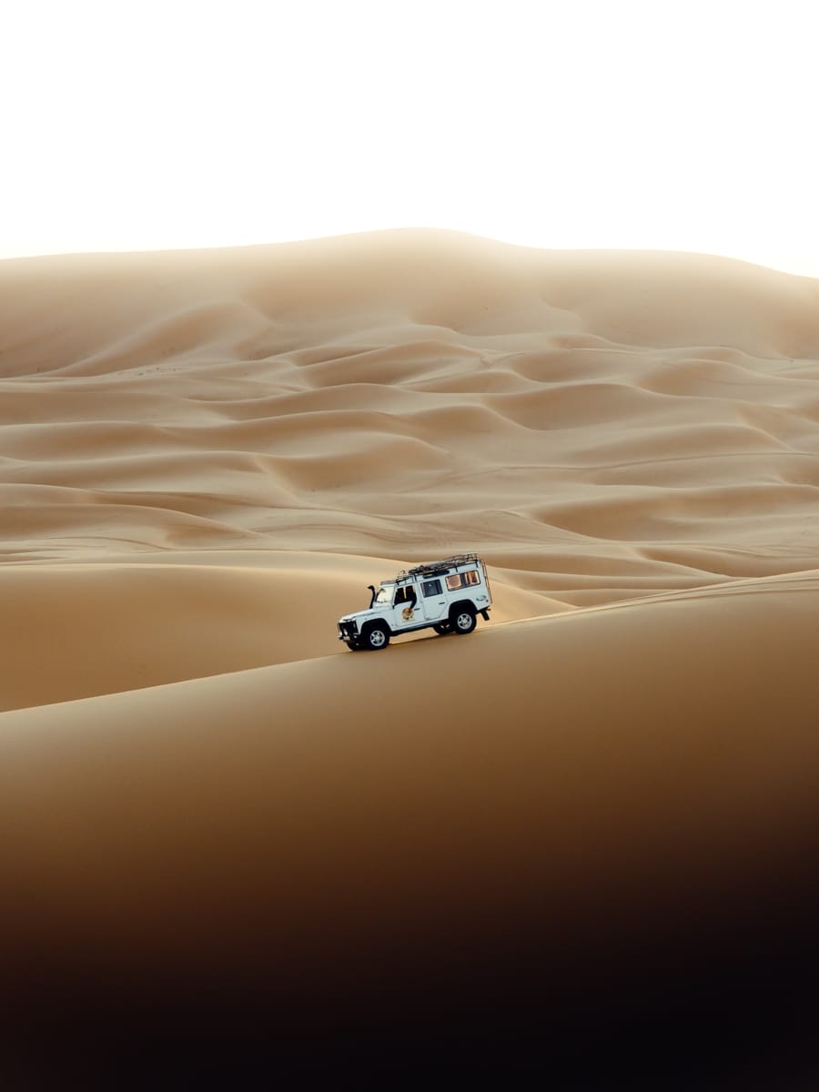 A white off-road vehicle drives up a steep sand dune, surrounded by vast desert landscapes under a bright sky—captured by Filippo Bellisola, an Italian content creator and outdoor filmmaker.