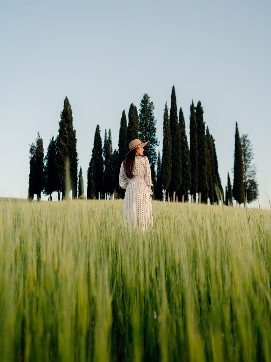 A Travel Content Creator in a long white dress and hat stands in a field of tall green grass, facing away, with a row of cypress trees in the background under a clear sky.