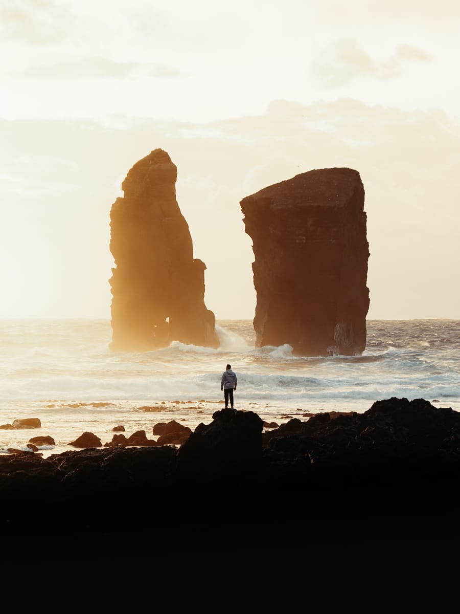 A person stands on a rocky shore, facing two large sea stacks rising from the sea, with mist and warm golden light in the background—captured by Filippo Bellisola, an Italian Content Creator and Outdoor Filmmaker.