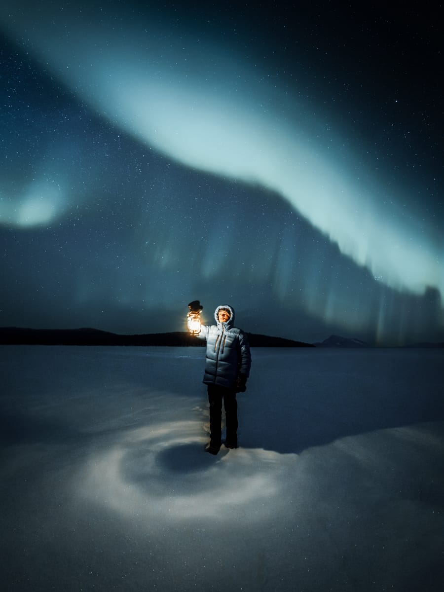 An outdoor content creator in a winter coat stands on snow at night, holding a lantern. The northern lights illuminate the sky above, creating a glowing, wavy pattern against the starry night.