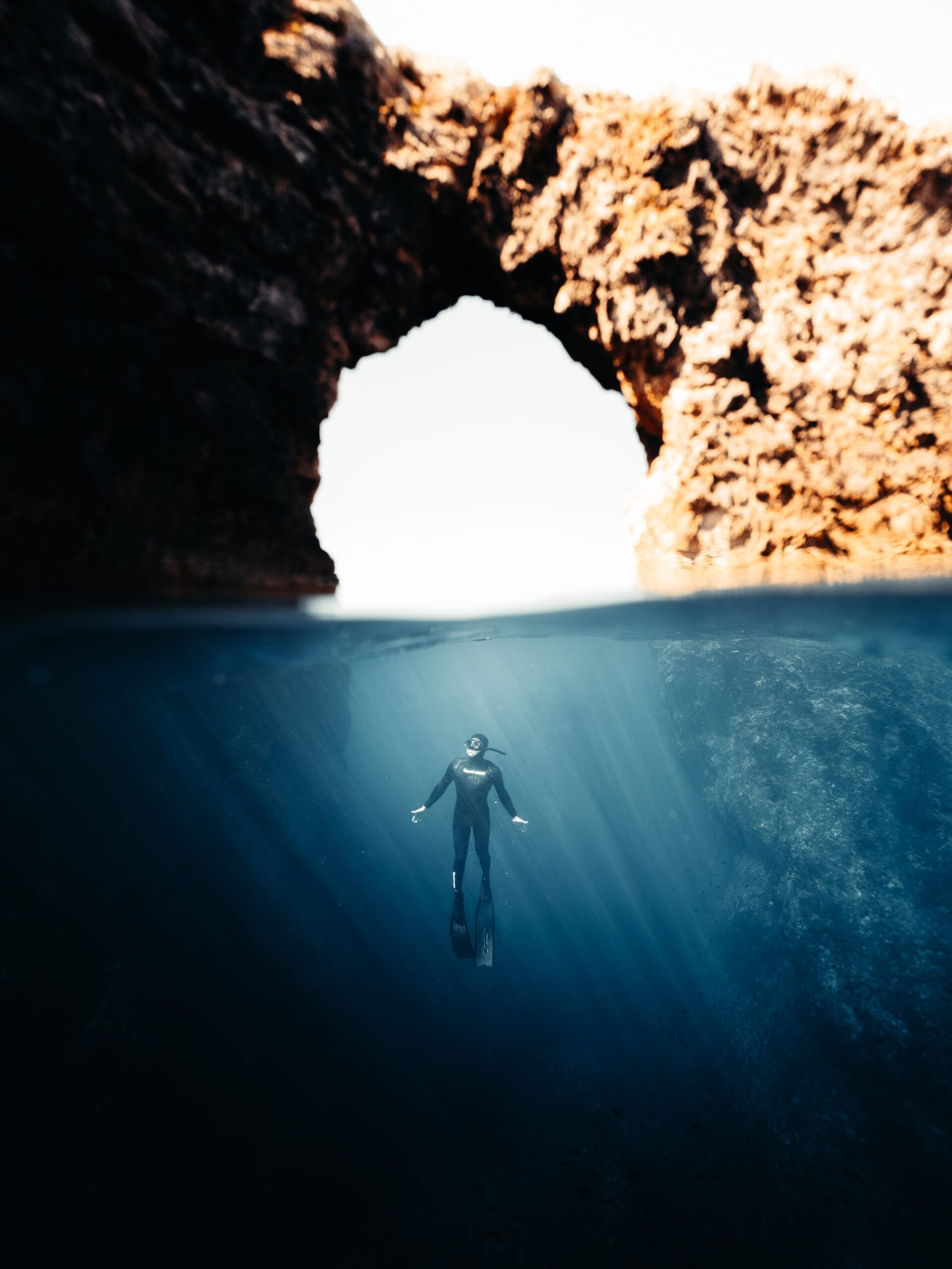 A Travel Content Creator in a wetsuit floats underwater near a rocky arch, with sunlight streaming down through clear blue water and illuminating the scene beneath the surface.