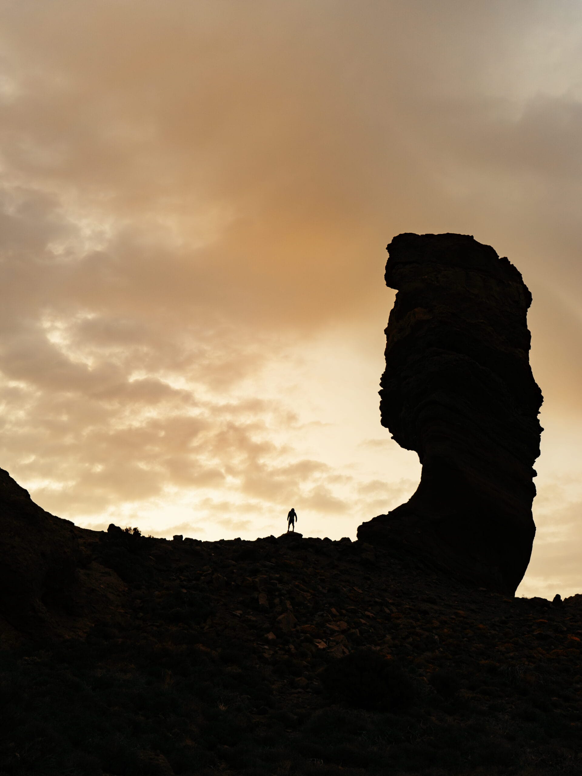 A silhouette of a person standing on rocky terrain beside a tall, uniquely shaped rock formation at sunset, with a dramatic, cloudy sky in the background—an inspiring scene perfect for adventurous brands.